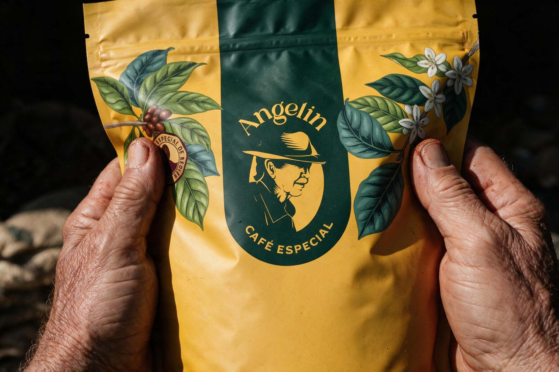 Mãos de produtor segurando o pouch amarelo de café especial Angelin, com ilustração de ramos de café e o selo da marca.